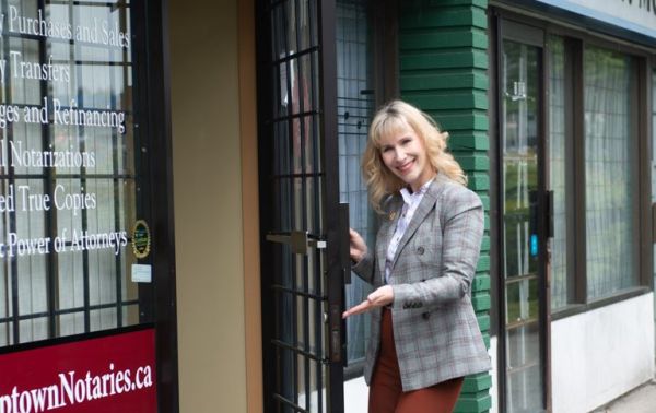 Smiling woman in a gray plaid blazer opens the door of a notary office storefront; service list and a notary sign are visible in the window.
