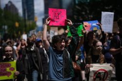Group of diverse protesters marching in a city street, with a bright pink sign showing 'AI' crossed out held high by a shouting participant in the foreground.