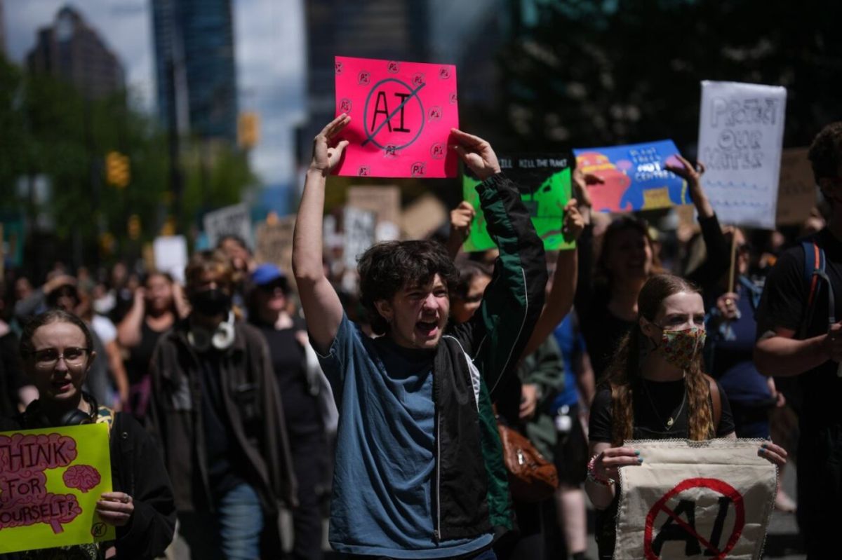 Большинство канадцев | Наш Ванкувер Group of diverse protesters marching in a city street, with a bright pink sign showing 'AI' crossed out held high by a shouting participant in the foreground.