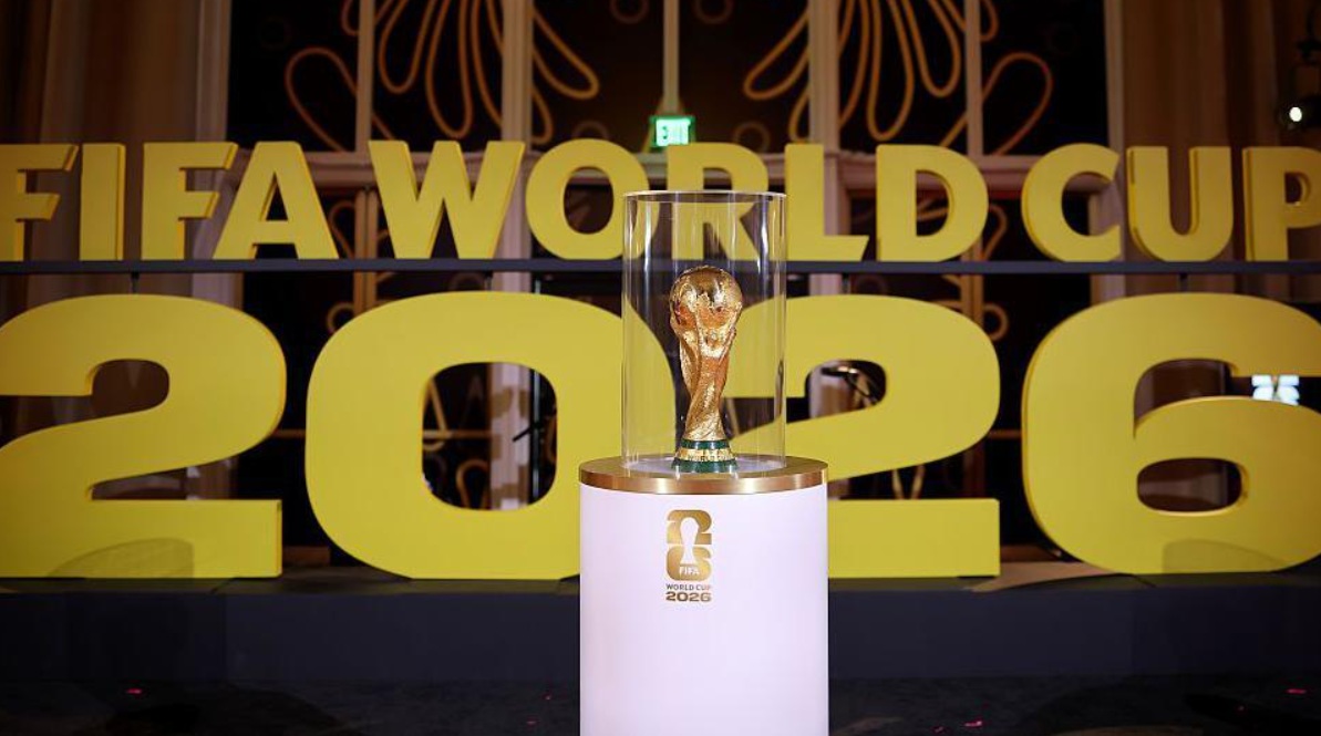 The FIFA World Cup 2026 trophy on a pedestal in a glass case, with oversized yellow '2026' and 'FIFA WORLD CUP' signage in the background.