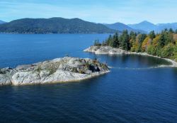 Rocky shoreline extending into a calm blue lake, with evergreen trees on a forested peninsula and distant mountains.