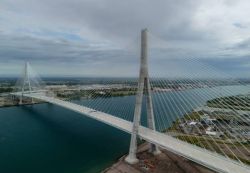Aerial view of a cable-stayed bridge with a tall central pylon and radiating white cables over blue water.