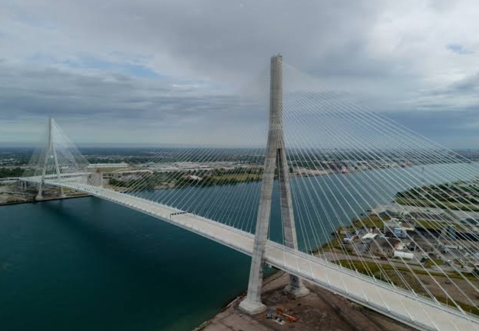 Aerial view of a cable-stayed bridge with a tall central pylon and radiating white cables over blue water.