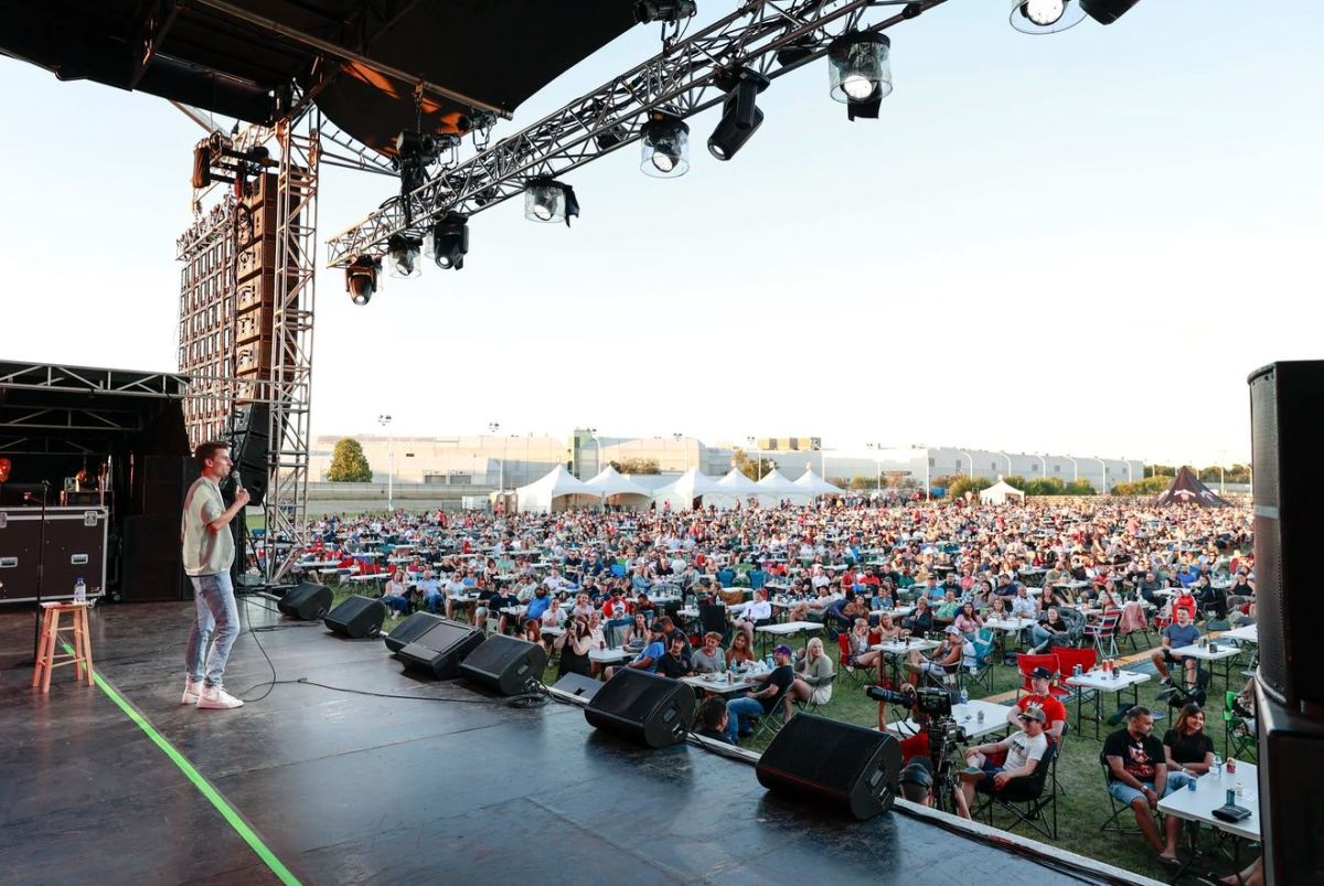 Speaker on an outdoor stage addressing a large festival crowd seated at tables and blankets afar.