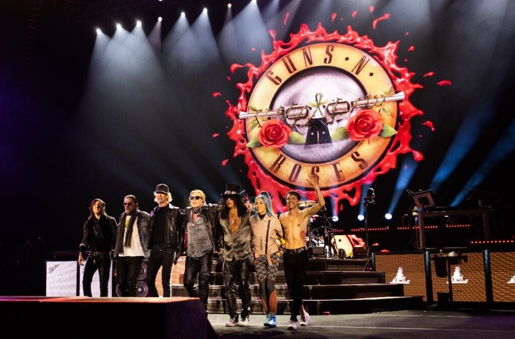 Concert scene: Guns N' Roses on stage with a large circular logo backdrop and red flames behind them, all members bowing or waving to the crowd.