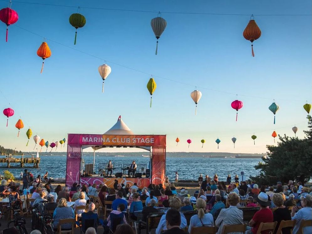 Outdoor concert by the water with a pink Marina Club Stage and a crowd watching performers, under colorful hanging lanterns by the shore.