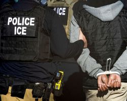 Police officer in tactical vest labeled POLICE ICE guiding a restrained detainee with hands cuffed behind their back