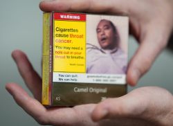 Hand holds a Camel Original cigarette pack with a bold yellow warning: 'Cigarettes cause throat cancer. You may need a hole cut in your throat to breathe.'