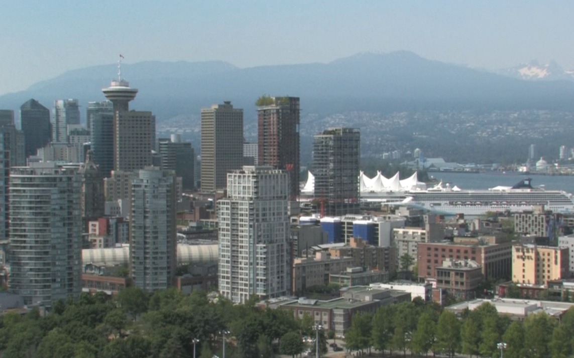 City skyline with tall modern buildings and a waterfront, mountains in the distance and white sail-like structures by the harbor (Canada Place).