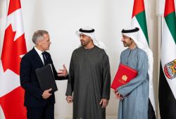 Three men in formal attire converse between flags of Canada and the UAE, each holding folders.