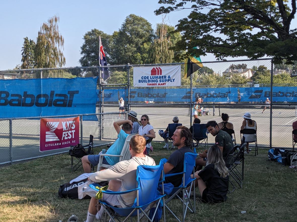 Spectators in folding chairs watching a tennis match along a chain-link fence, with blue Babolat banners and sponsor signs nearby on a sunny day.