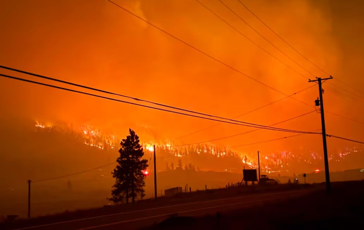 Wildfire consumes hillside at dusk with an orange sky; power lines cross the frame and a road runs foreground.