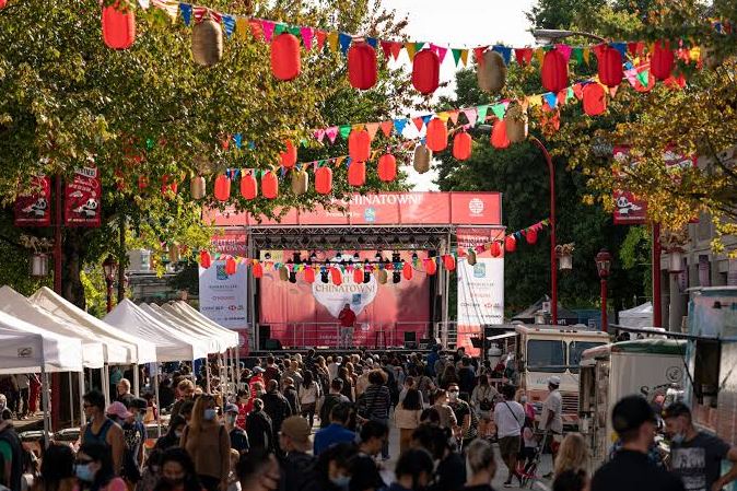 Crowd at an outdoor festival with a stage and red lantern bunting across the street, tents lining the sides, and a banner reading Chinatown on stage.