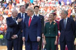 Group of formally dressed officials on a stage at an outdoor ceremony, with a large crowd in the stands behind them.