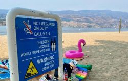 Blue beach safety sign warns no lifeguard on duty and to call 9-1-1; children require adult supervision and there is a sudden drop-off warning, with a pink flamingo pool float nearby and beach gear on the sand.
