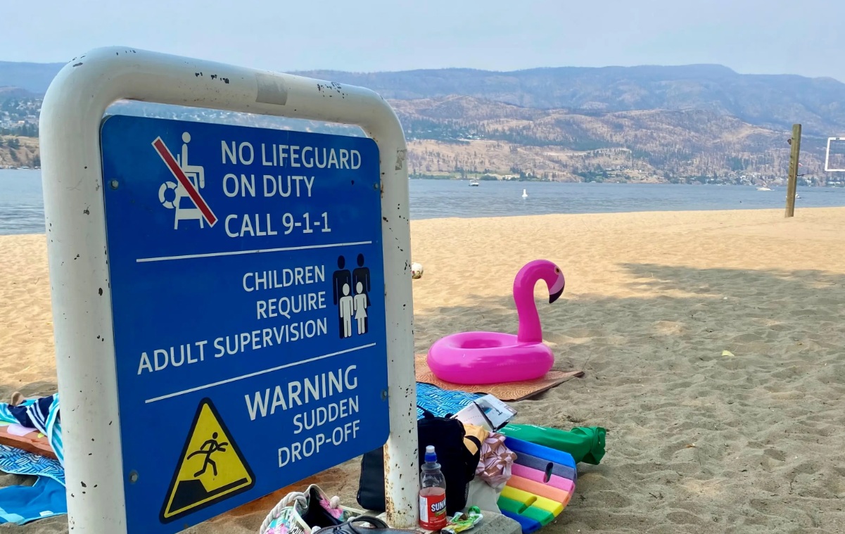 Blue beach safety sign warns no lifeguard on duty and to call 9-1-1; children require adult supervision and there is a sudden drop-off warning, with a pink flamingo pool float nearby and beach gear on the sand.