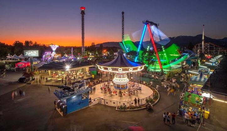 Outdoor amusement park at sunset featuring a lit carousel in the center, a neon roller coaster, and visitors wandering among stalls and rides.