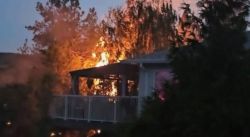 Deck or balcony of a house engulfed in flames with trees in the background at dusk/early evening.
