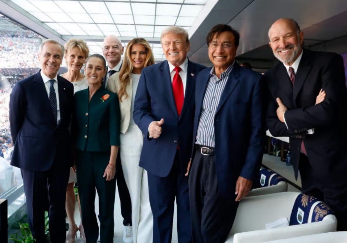 Group of well-dressed people posing for a photo in a stadium luxury box, smiling and giving thumbs up wrist to camera.