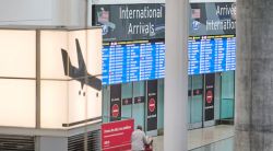 Airport arrivals area with large 'International Arrivals' signs and blue flight information boards above glass doors.