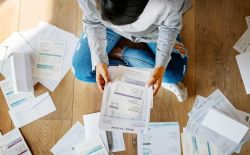 Person sits on a wooden floor, surrounded by scattered invoices and bills, holding one invoice in their hands.