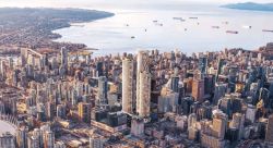 A dense city skyline with two tall modern towers and a harbor with numerous ships beyond the buildings, viewed from above.