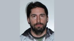 Portrait of a man with dark hair and a beard, wearing a light hoodie, looking at the camera.