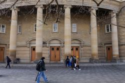 Stone façade with large columns and wooden doors, pedestrians walking across a cobblestone plaza in front of a historic building.