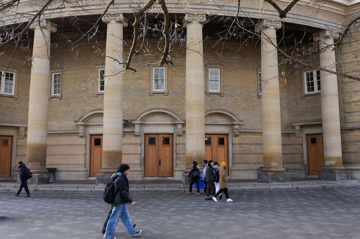 Stone façade with large columns and wooden doors, pedestrians walking across a cobblestone plaza in front of a historic building.