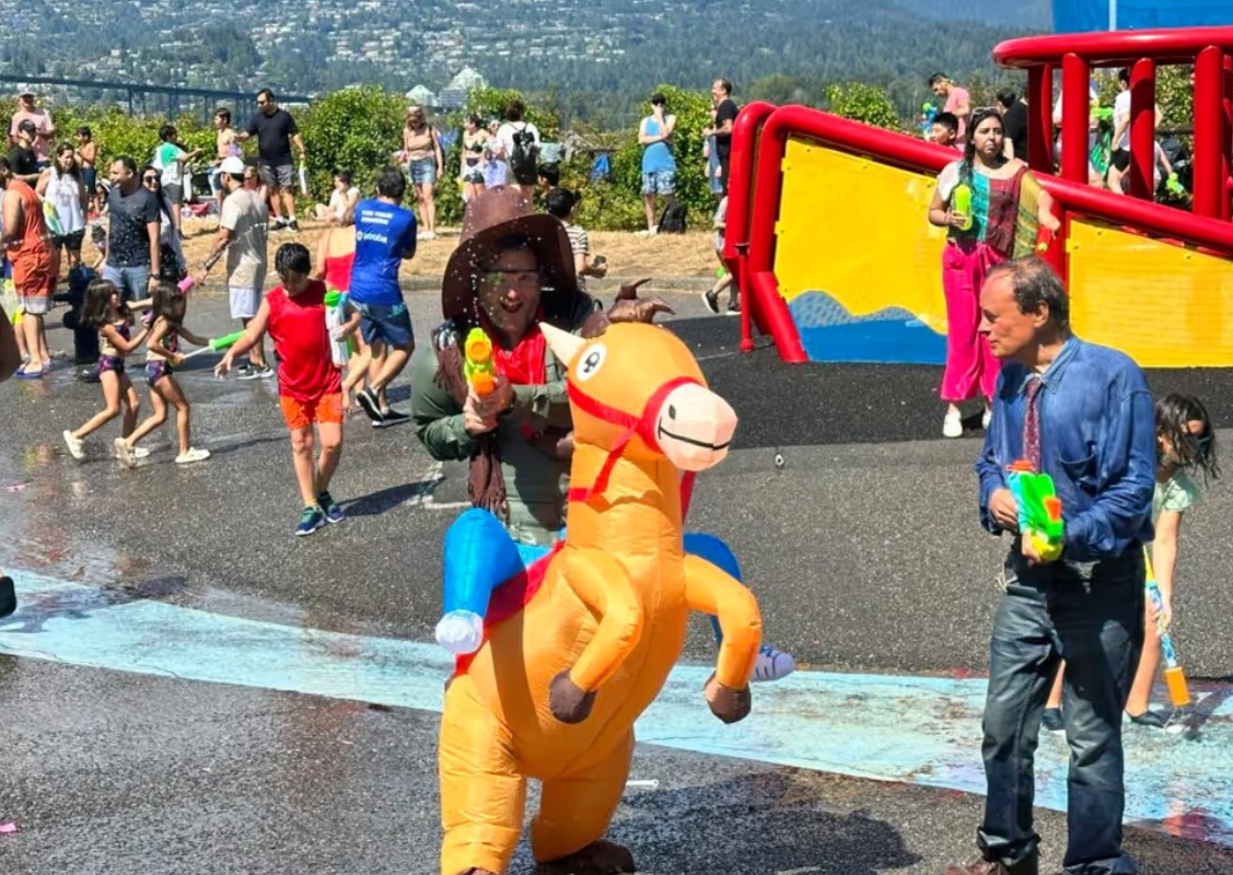 People enjoy a sunny water park; an inflatable orange dinosaur rider moves along as others spray water nearby, with a bright red-yellow slide in the background and mountains beyond.