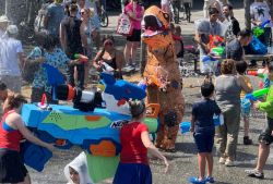 Group of people spraying water guns at a festival; a large blue Nerf gun dominates the foreground while a person in a dinosaur costume stands nearby.