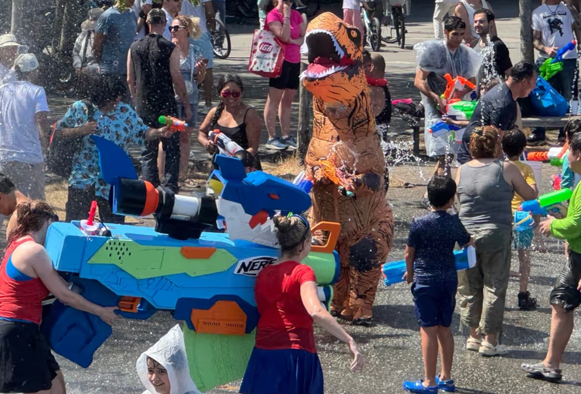 Group of people spraying water guns at a festival; a large blue Nerf gun dominates the foreground while a person in a dinosaur costume stands nearby.