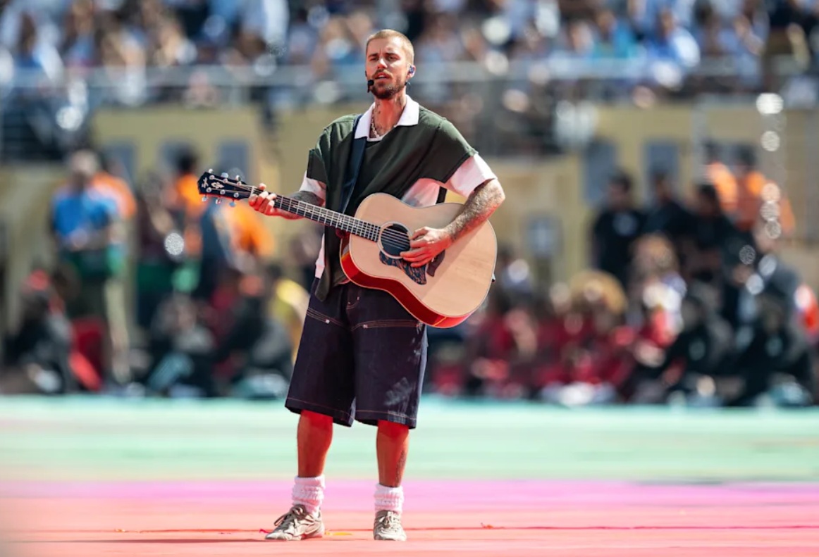 Male street-musician playing an acoustic guitar on a colorful outdoor stage with a cheering crowd in the background