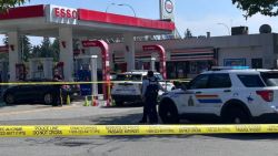 Gas station scene with Esso branding, police cars, and yellow crime-scene tape blocking the area.