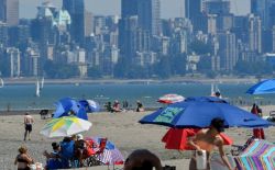 People relaxing on a crowded beach with colorful umbrellas and a distant city skyline across the water.