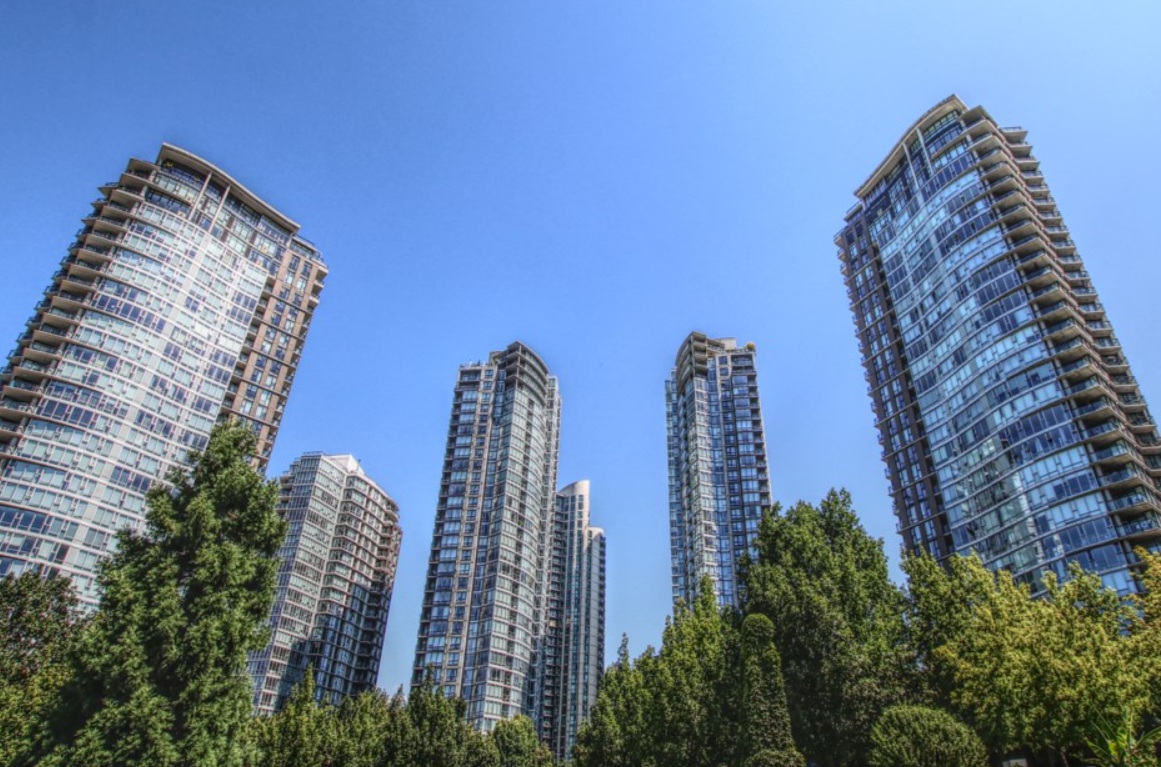 Tall glass apartment towers rise above green trees under a clear blue sky.