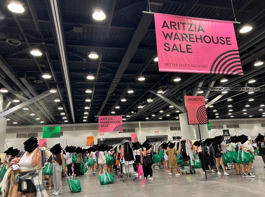 Interior of a large sale event with pink banners reading 'ARITZIA WAREHOUSE SALE'; clothing racks fill the space and shoppers with green bags browse the aisles.