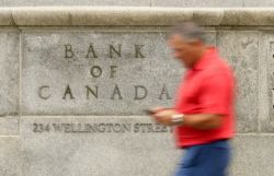 Blurry man in a red shirt walking past a Bank of Canada sign engraved in a stone wall.
