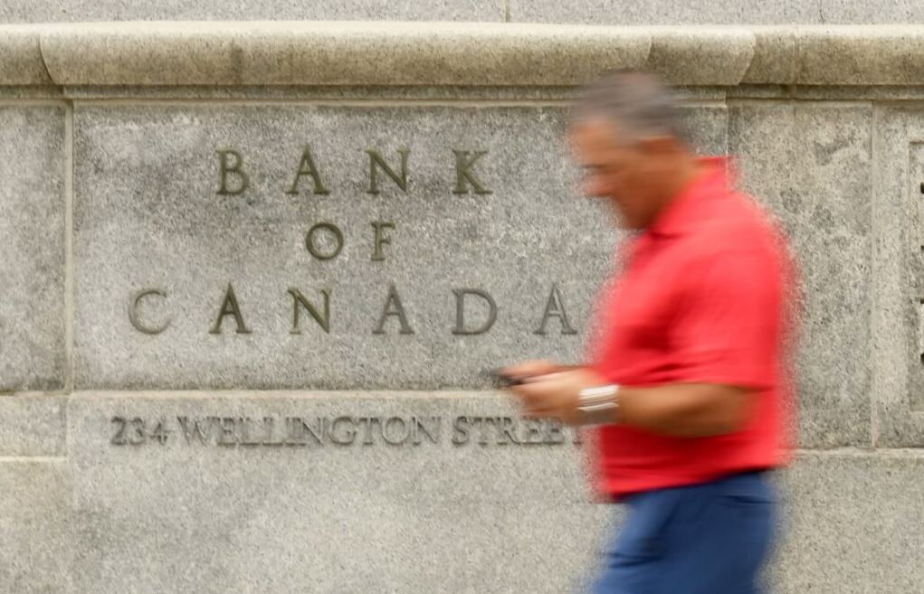 Blurry man in a red shirt walking past a Bank of Canada sign engraved in a stone wall.