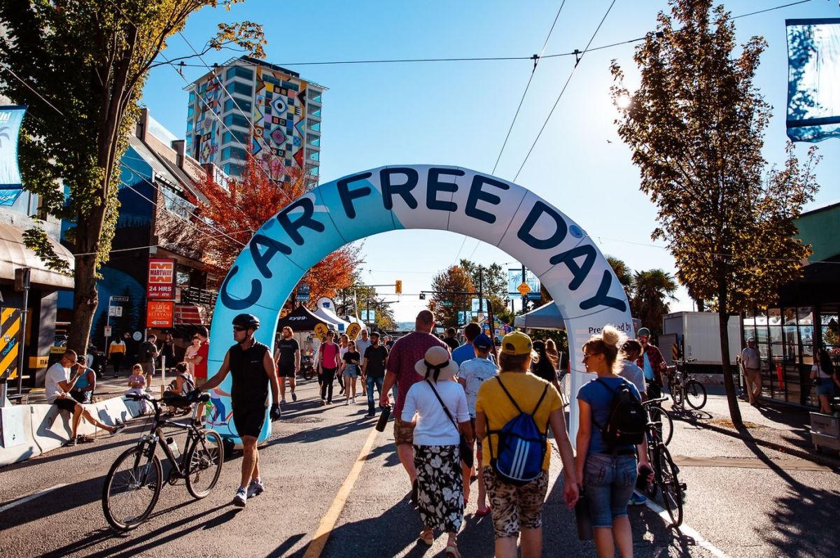 People walk and bike under a large inflatable arch that reads 'CAR FREE DAY' on a sunny street festival.