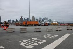 Port/harbor loading area with an orange Hapag-Lloyd truck, pallets, and containers, and a city skyline in the background.