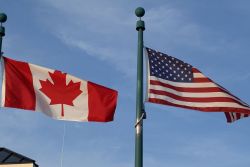 Canadian flag and American flag waving on adjacent flagpoles against a clear blue sky.