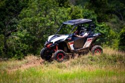 Off-road side-by-side UTV with a helmeted driver cresting a grassy hill, forest in the background.