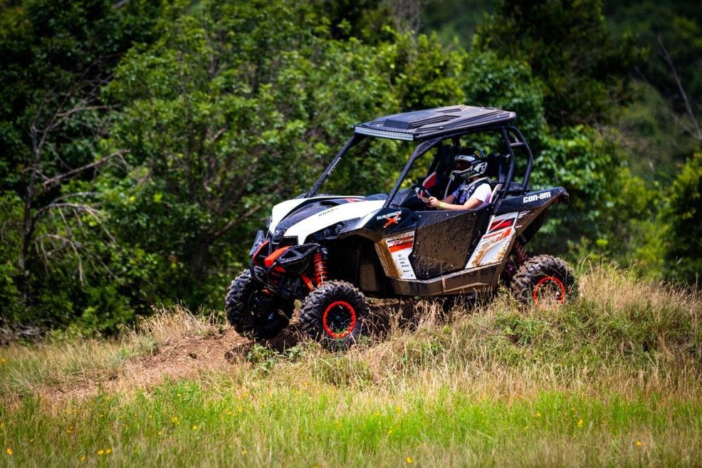 Off-road side-by-side UTV with a helmeted driver cresting a grassy hill, forest in the background.