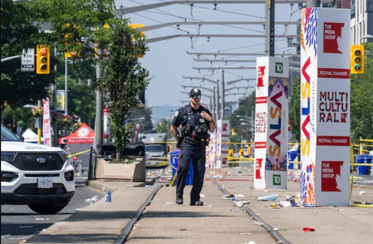 Uniformed police officer standing in the middle of a littered urban street, with a car on the left and festival banners ahead.