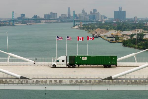 White semi-truck pulls a green shipping container across a suspension bridge, with US and Canadian flags and a distant city skyline in the background.