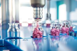 Automated pill-filling line placing pink tablets into glass vials on a blue conveyor.