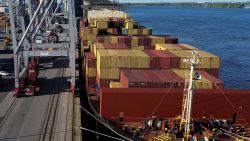 Container ship with stacked colorful cargo containers beside a pier and blue water, cranes on the left in a busy port. This shows a loading dock scene at sea.