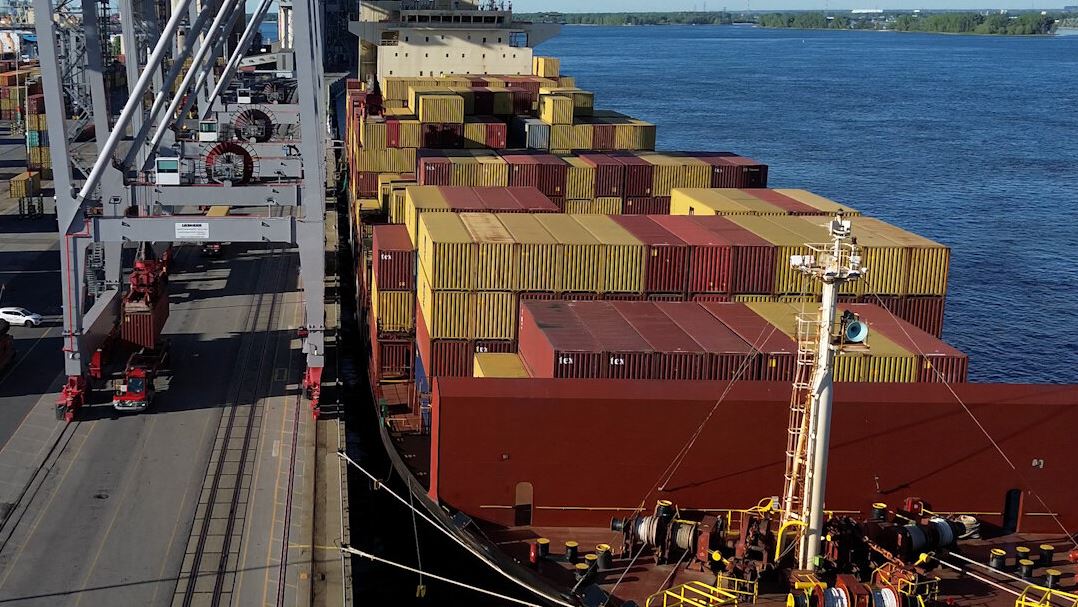 Container ship with stacked colorful cargo containers beside a pier and blue water, cranes on the left in a busy port. This shows a loading dock scene at sea.