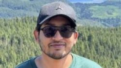 Man wearing a dark cap and sunglasses outdoors with a forested mountainside behind him